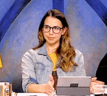 a woman wearing glasses and a denim jacket is sitting at a table with a tablet