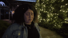 a woman in a denim jacket is standing in front of a christmas tree and says you