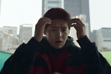 a man in a black and red jacket has his hands on his forehead in front of a city skyline
