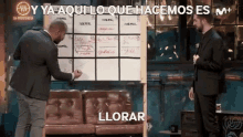 a man in a suit is writing on a white board next to a couch while another man looks on and says llorar