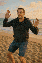 a man wearing glasses and a black hoodie stands on the beach
