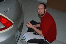 a man in a red sweater sits in front of a silver car with the letters su on the back
