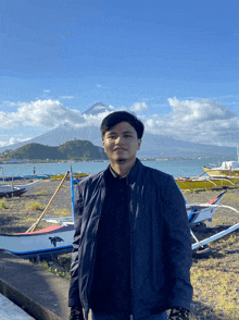 a man in a black jacket stands in front of boats on the beach