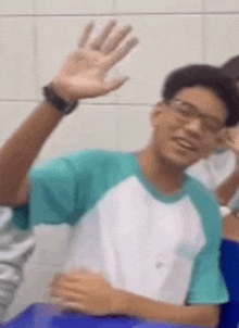 a young man is waving his hand in a classroom while wearing glasses and a watch .