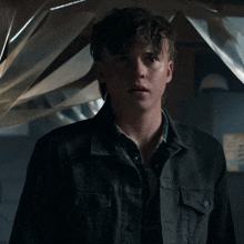 a young man wearing a black jacket and a plaid shirt stands in a dark room