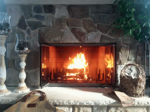 A gif of a blazing fire in a stone heart fireplace. In front of the fireplace there's a fancy wicker basket and some tall vases with glass bowls filled with rocks or something. It's classic cottagecore bougie country estate - chic.