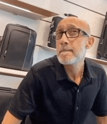 a bald man with glasses and a beard is sitting in front of a display of luggage