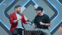 a man in a red jacket is talking into a microphone next to another man in a black shirt who is sitting down