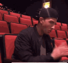 a man in a black jacket and hat is sitting in an auditorium .