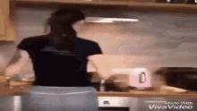 a woman in a black shirt is standing in a kitchen with a pink kettle .