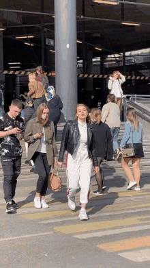a woman in a leather jacket is walking across a street while a group of people look on