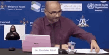 a man in a red shirt and tie is sitting at a desk with a laptop .