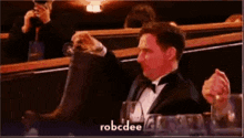 a man in a tuxedo is sitting at a table with a glass of wine and a bottle of water .