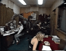 a group of people are gathered in a room including a woman sitting at a desk