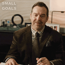 a man in a suit and tie sits at a desk with a small achievable goals advertisement behind him