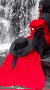 a woman in a red dress is sitting in front of a waterfall wearing a black hat