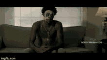 a shirtless man with a clown mask on his face is sitting on a couch ..