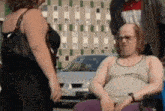 a man in a tank top and glasses is sitting in a wheelchair while a woman walks behind him .