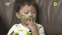 a little boy with green paint on his face is sitting on a couch eating a cucumber .