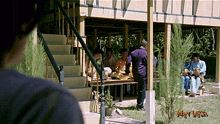 a man is looking at a group of people sitting under a porch with the words wan laksa on the bottom