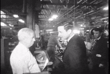 a man in a suit talks to a man in a white shirt in a factory