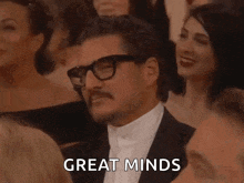 a man in a tuxedo with glasses and a mustache is sitting in a crowd of people .