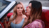 two women are sitting in a red convertible car and one of them is holding her finger to her lips .