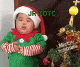 a baby dressed as an elf is standing in front of a christmas tree and a sign that says merry christmas .