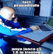 a bald man is sitting at a table with a laptop and talking on a cell phone .
