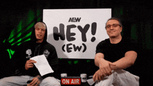 two men sitting in front of a sign that says hey