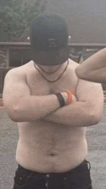 a shirtless man wearing a hat with the letter r on it is standing with his arms crossed