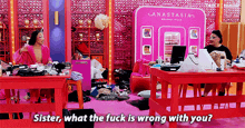 two women are sitting at red tables in front of a pink wall and talking .