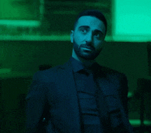 a man in a suit and tie stands in a dark room with a green light behind him
