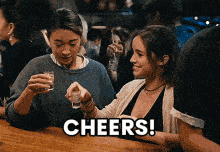 two women are sitting at a bar holding shot glasses and the word cheers is on the bottom of the image