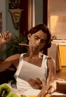 a man in a white tank top is smoking a cigarette while sitting at a table with a piece of paper .