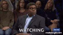a man in a suit and glasses is sitting in front of a crowd of people and watching .