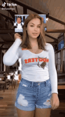 a woman wearing a hooters shirt and shorts is standing in front of a tv .