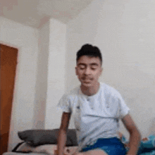 a young man in a white shirt and blue shorts is sitting on a bed and making a funny face .