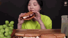 a woman in a green shirt is eating a waffle sandwich with grapes in the background