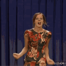 a woman in a floral dress is standing in front of a blue curtain .