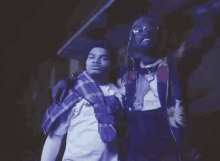 two men are posing for a picture together in a dark room . one of the men is wearing a plaid shirt .