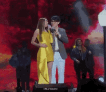 a woman in a yellow dress stands next to a man holding a microphone with the word mayward on the bottom