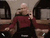 a bald man in a red shirt is sitting in a chair with the words engage below him