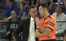 a man in an orange shirt with the word medicana on it talks to another man in a suit