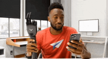 a man wearing a red shirt with a paper airplane on it is holding a phone and a prosthetic hand