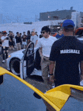 a man wearing a shirt that says marshall stands in front of a yellow car
