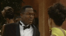 a man in a tuxedo and bow tie talking to a woman in a yellow dress .