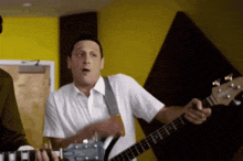 a man in a white shirt is playing a guitar in front of a yellow wall