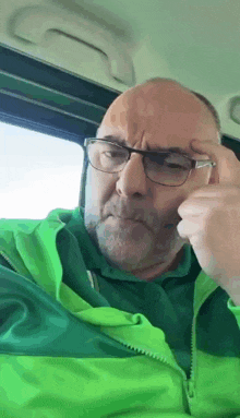 a man wearing glasses and a green jacket is scratching his forehead