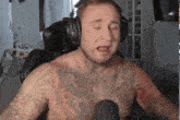 a shirtless man with tattoos is wearing headphones and singing into a microphone while sitting in a chair .